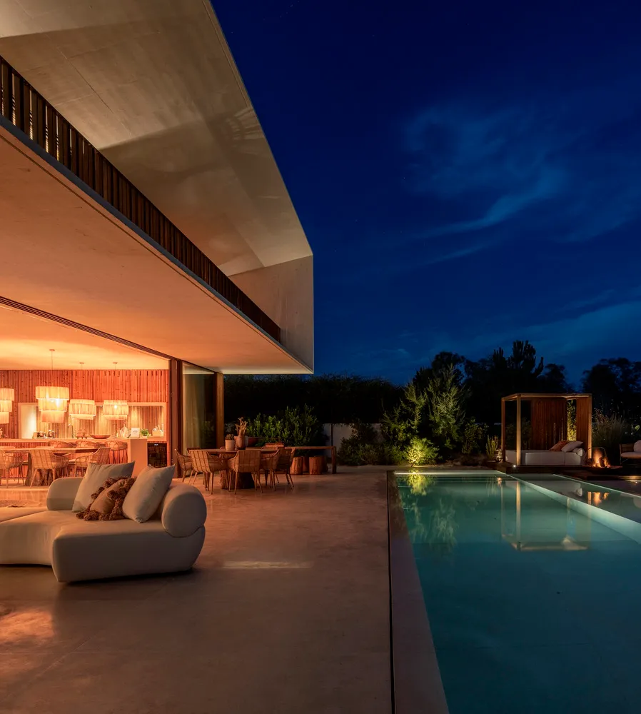 A modern residential patio with minimalist furniture and a large swimming pool under a dark, starry sky.