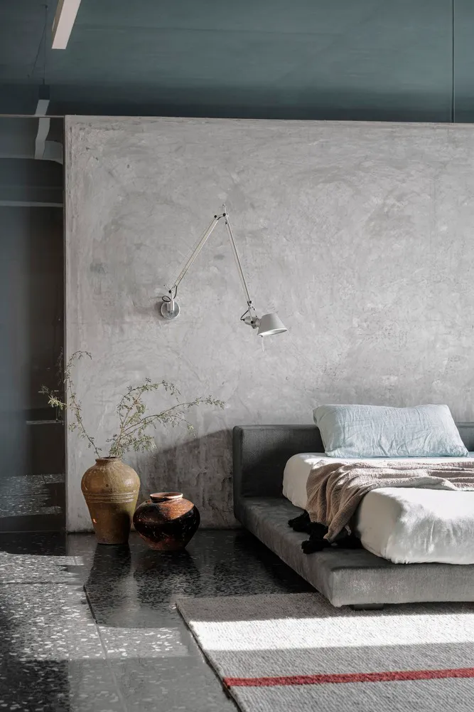 A minimalist bedroom with a concrete wall, a bed, a wall lamp, and decorative vases.