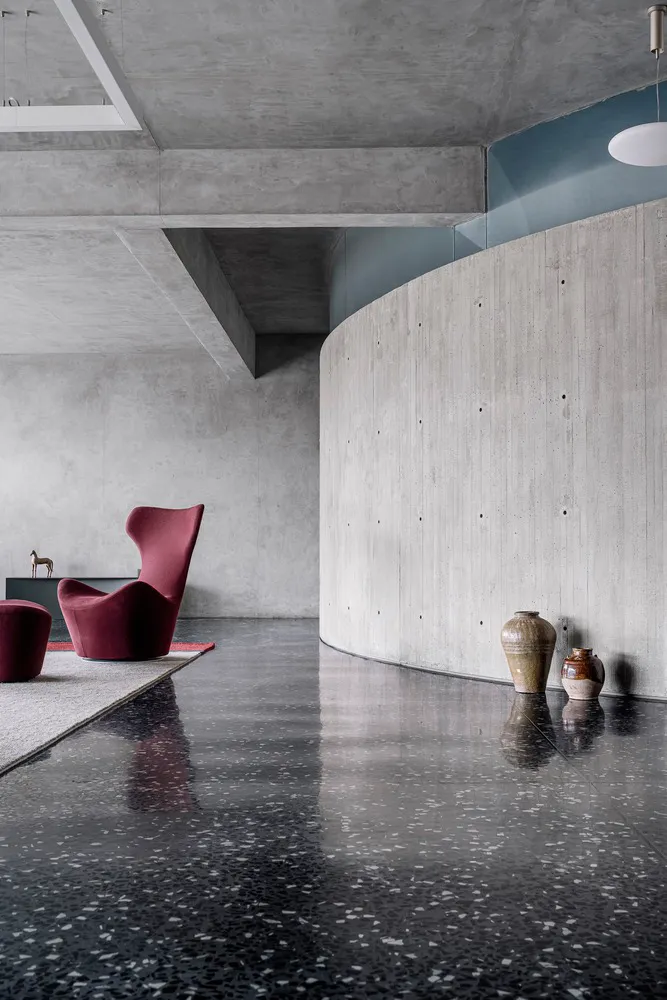 A minimalist living room with concrete walls, a red armchair, and terrazzo flooring.