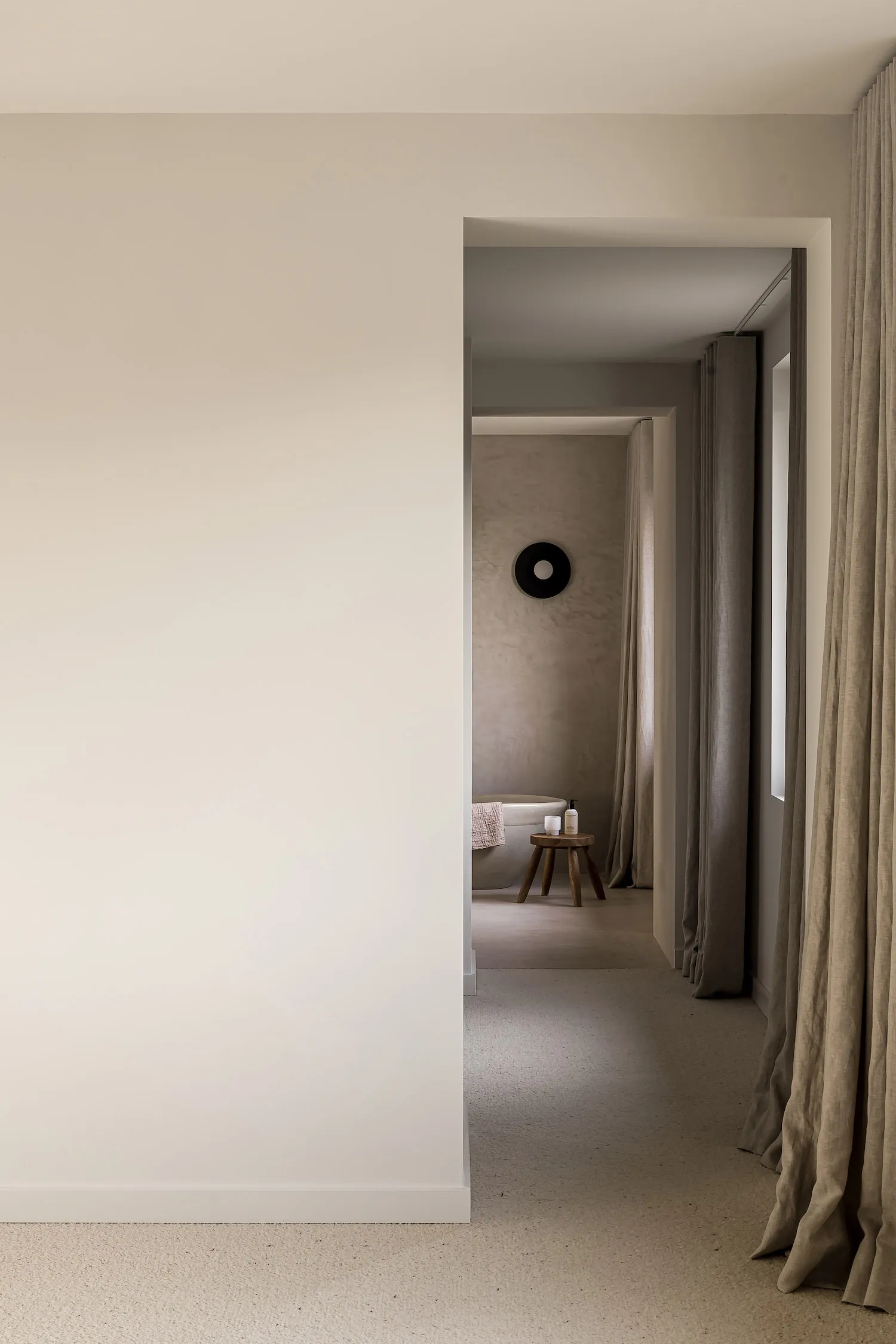 A minimalist bedroom with neutral tones, featuring a white wall, beige curtains, and a simple wooden stool.