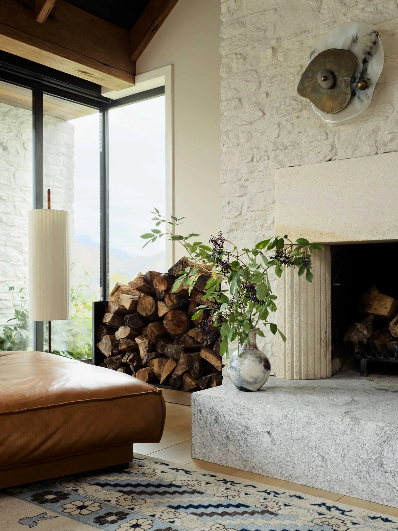 A stone fireplace in a living room with wood logs and a vase of greenery on the mantle.