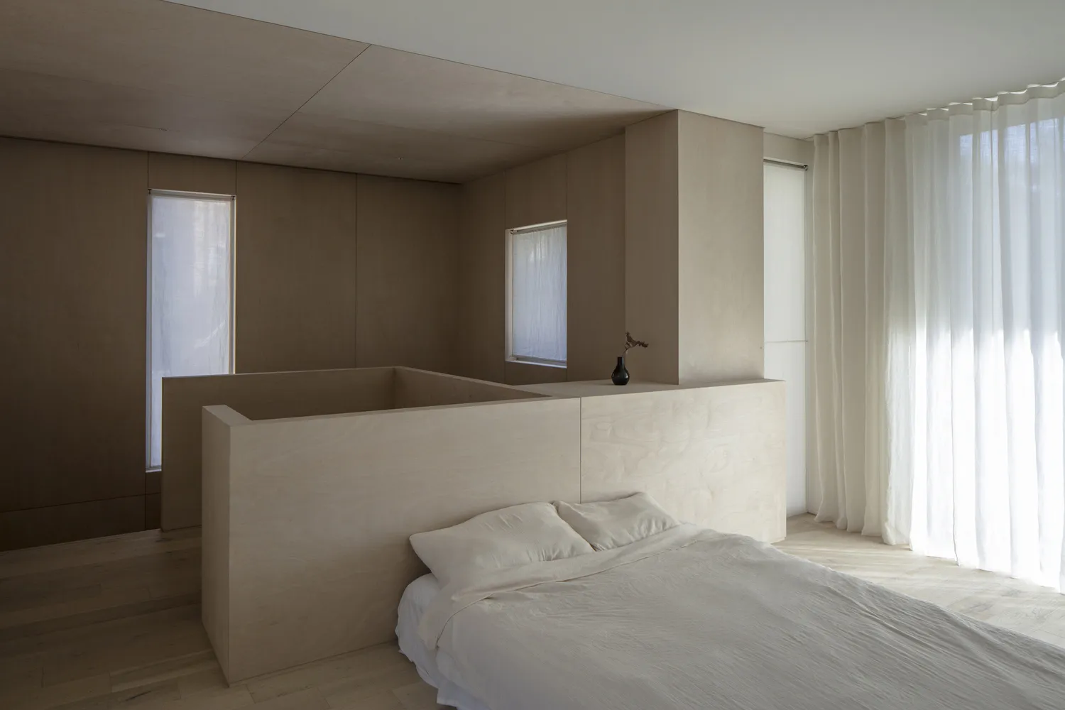 A sleek, contemporary bedroom with minimalist wooden paneling and a bed, featuring white bedding and sheer curtains.