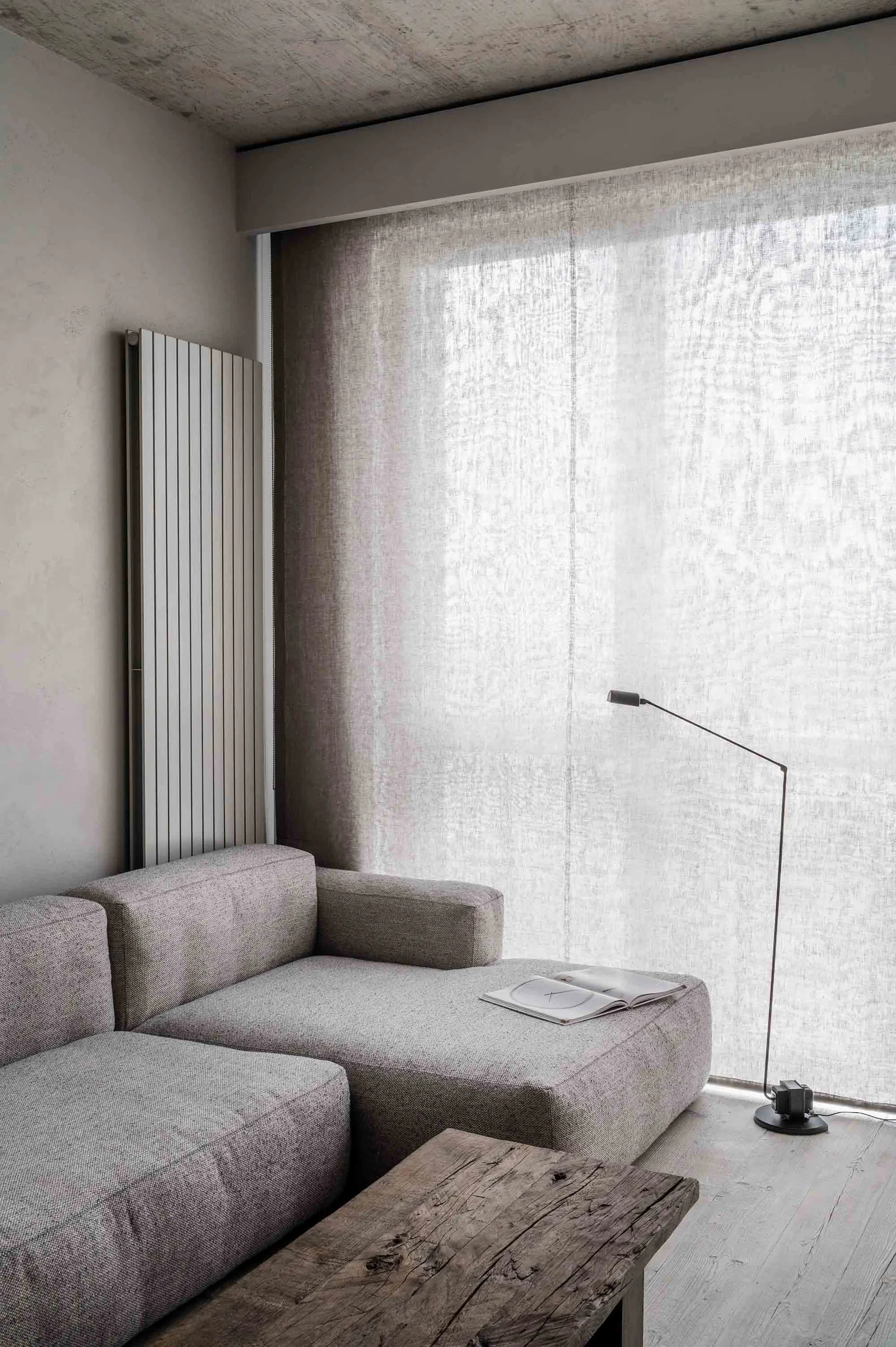 A minimalist living room with a light-colored fabric sofa, concrete radiator, cotton curtains, and a wooden coffee table.