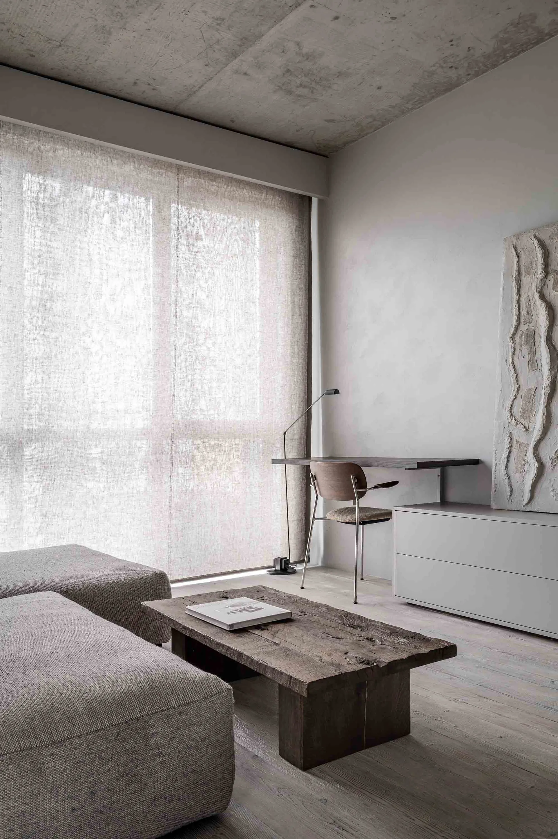 A modern minimalist living room with concrete walls, a grey sectional sofa, and a wooden coffee table.