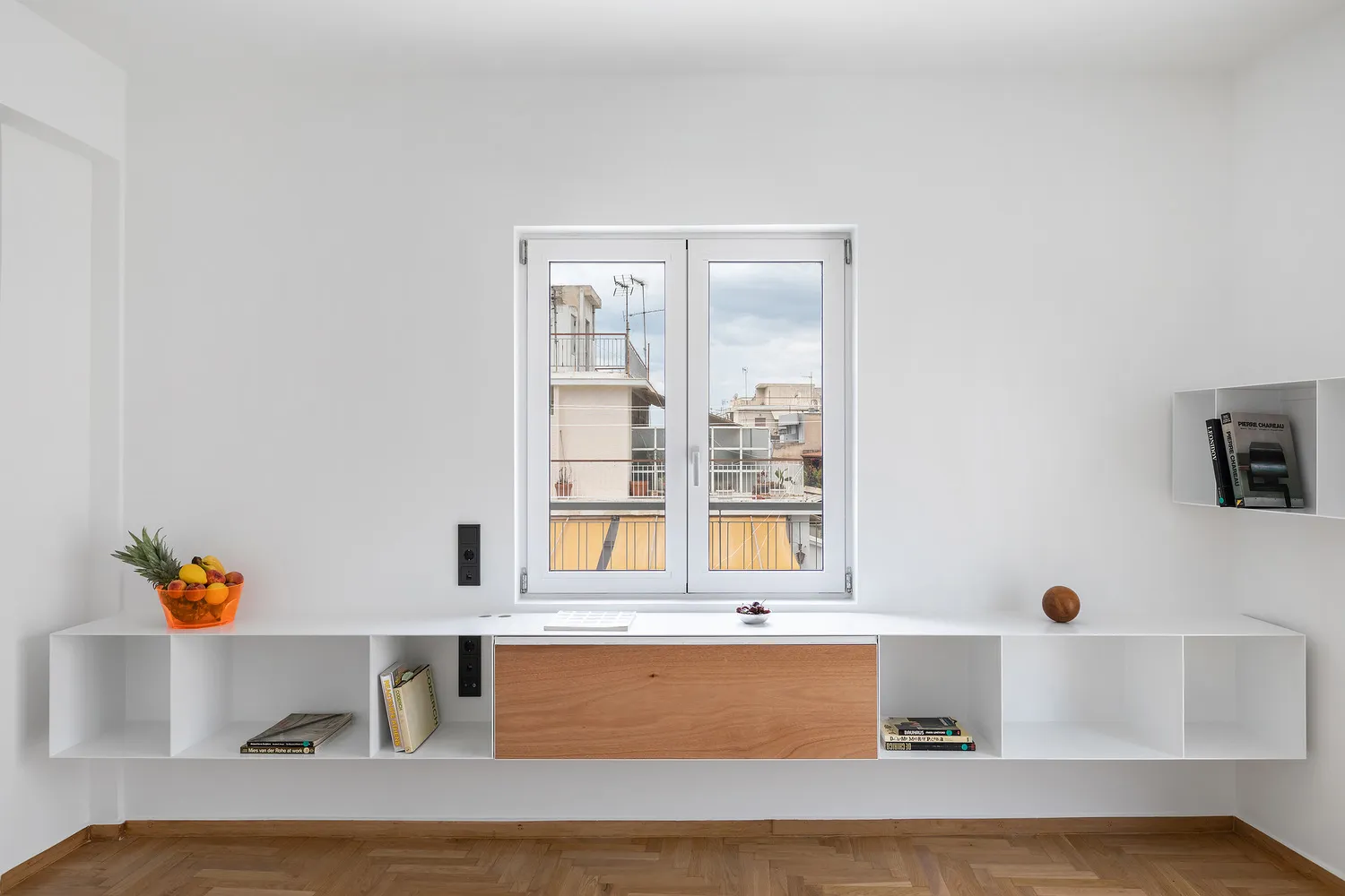 A minimalist residential interior featuring white walls, a large open window with a view of neighboring buildings, and wooden