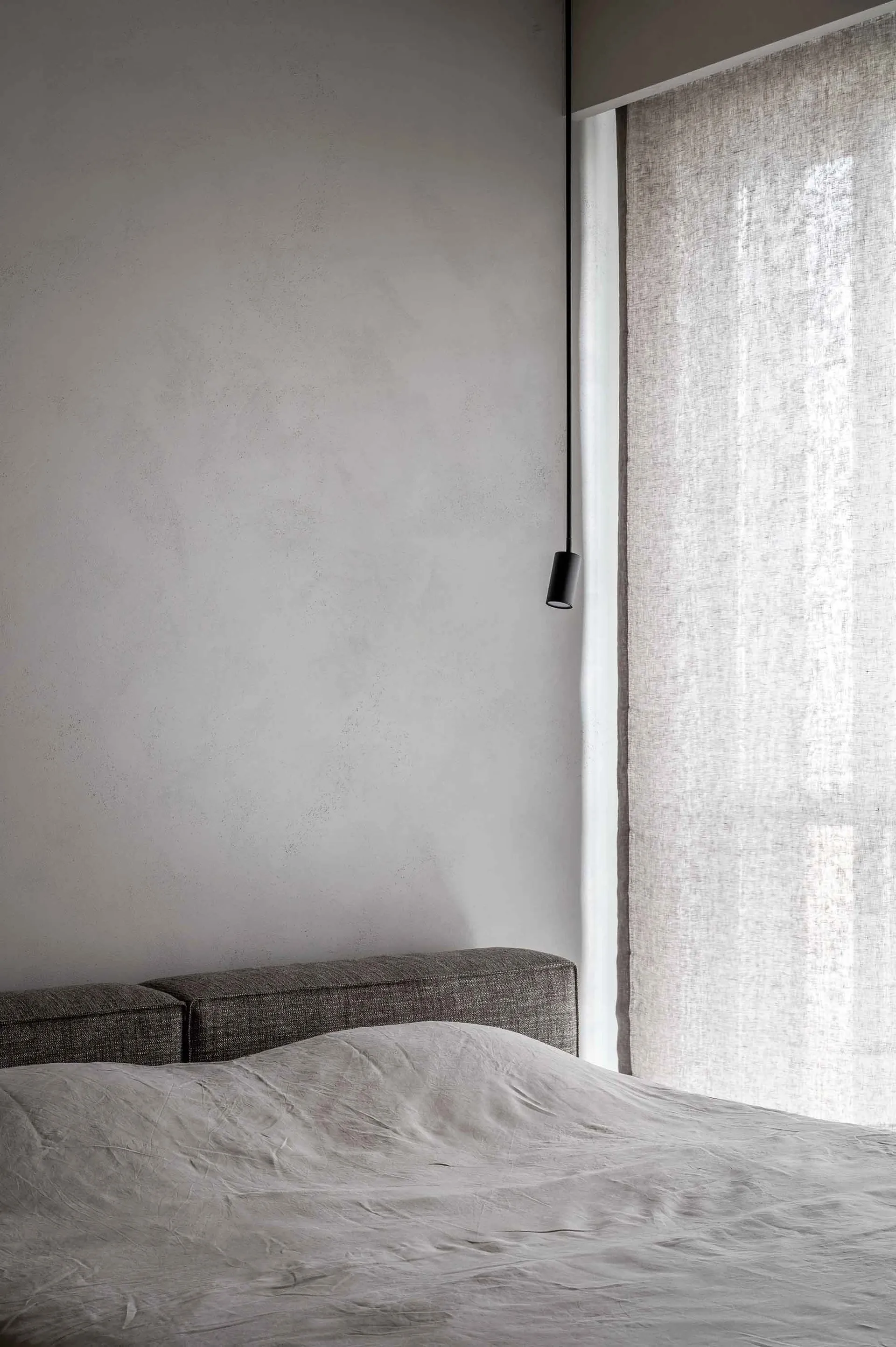 A modern grey bedroom featuring a bed draped in light grey fabric, minimalistic design, and natural light filtering through s
