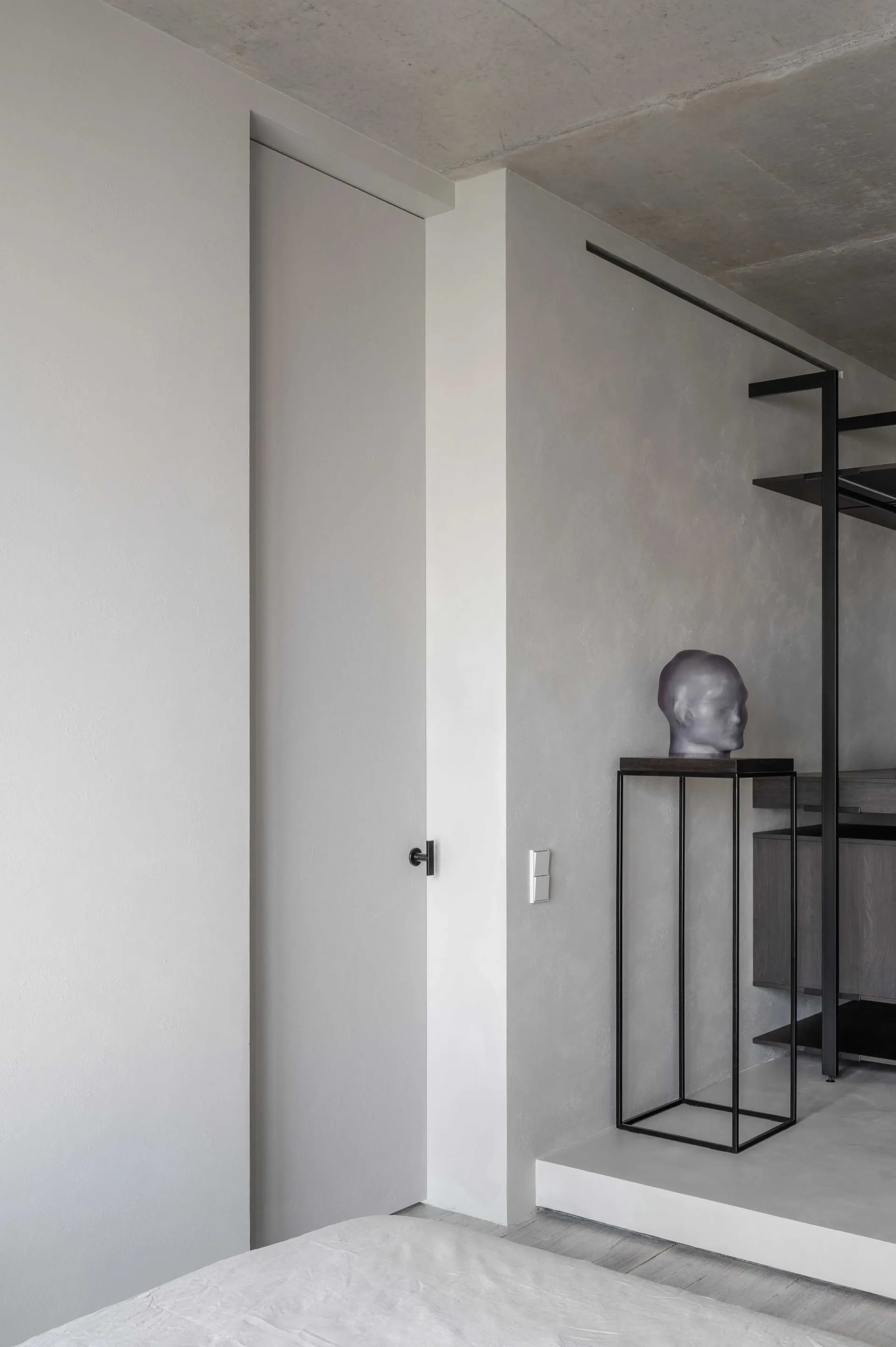 A minimalist bedroom featuring a concrete wall and floor, with a bed and a sculpture on a pedestal.