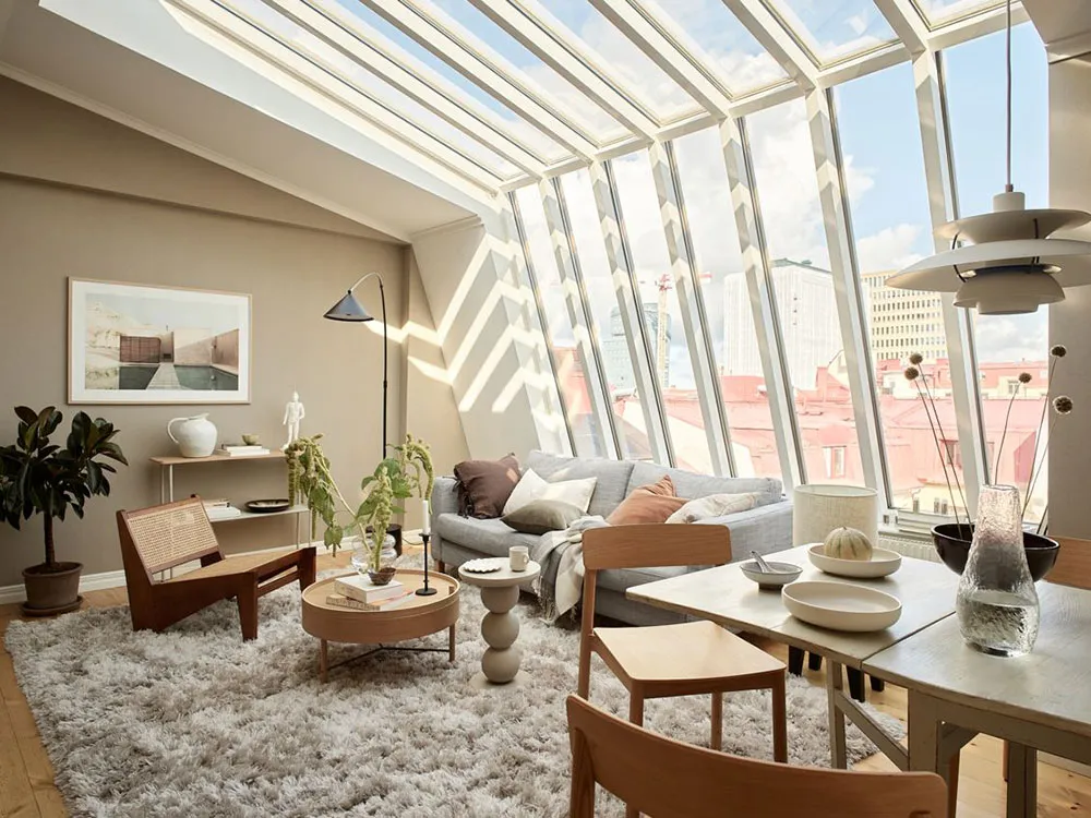 A minimalist living room with a grey sofa, wooden chairs, and a large window letting in natural light.