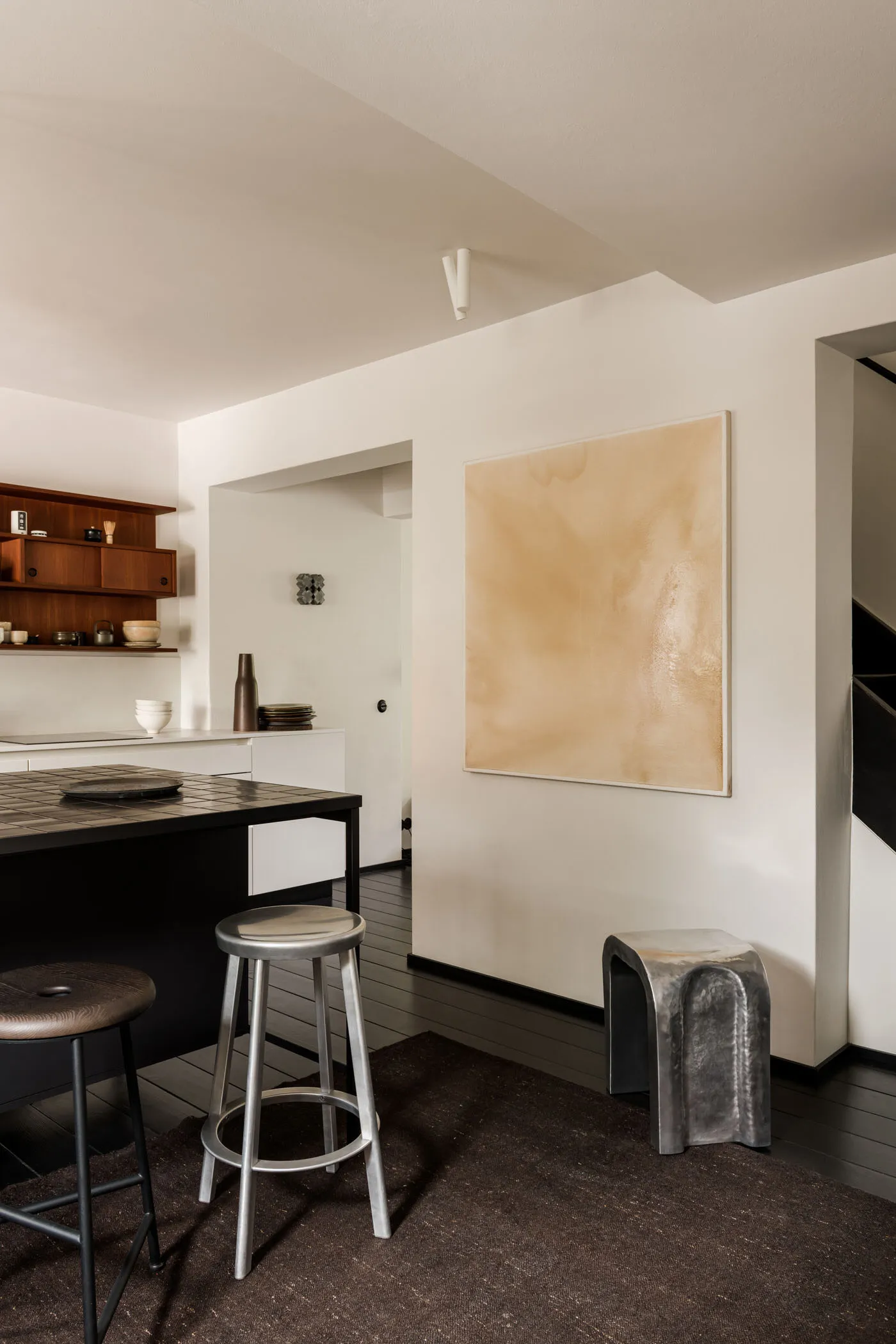 A minimalist residential kitchen featuring metal stools, wooden floor, and a large beige abstract painting on the wall.