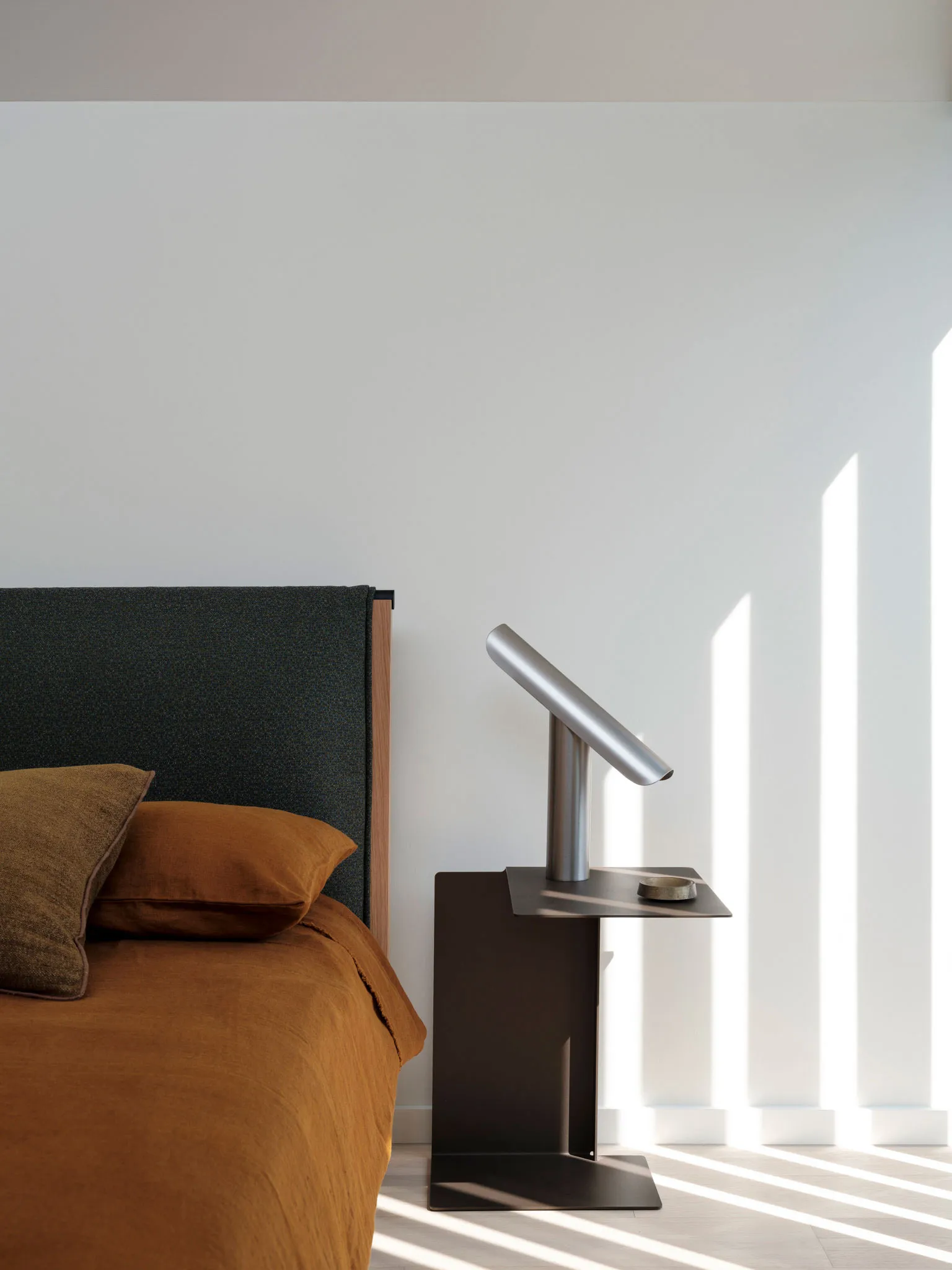 A minimalistic bedroom with a grey upholstered bed headboard and orange bedding, featuring a sleek, metallic bedside lamp wit