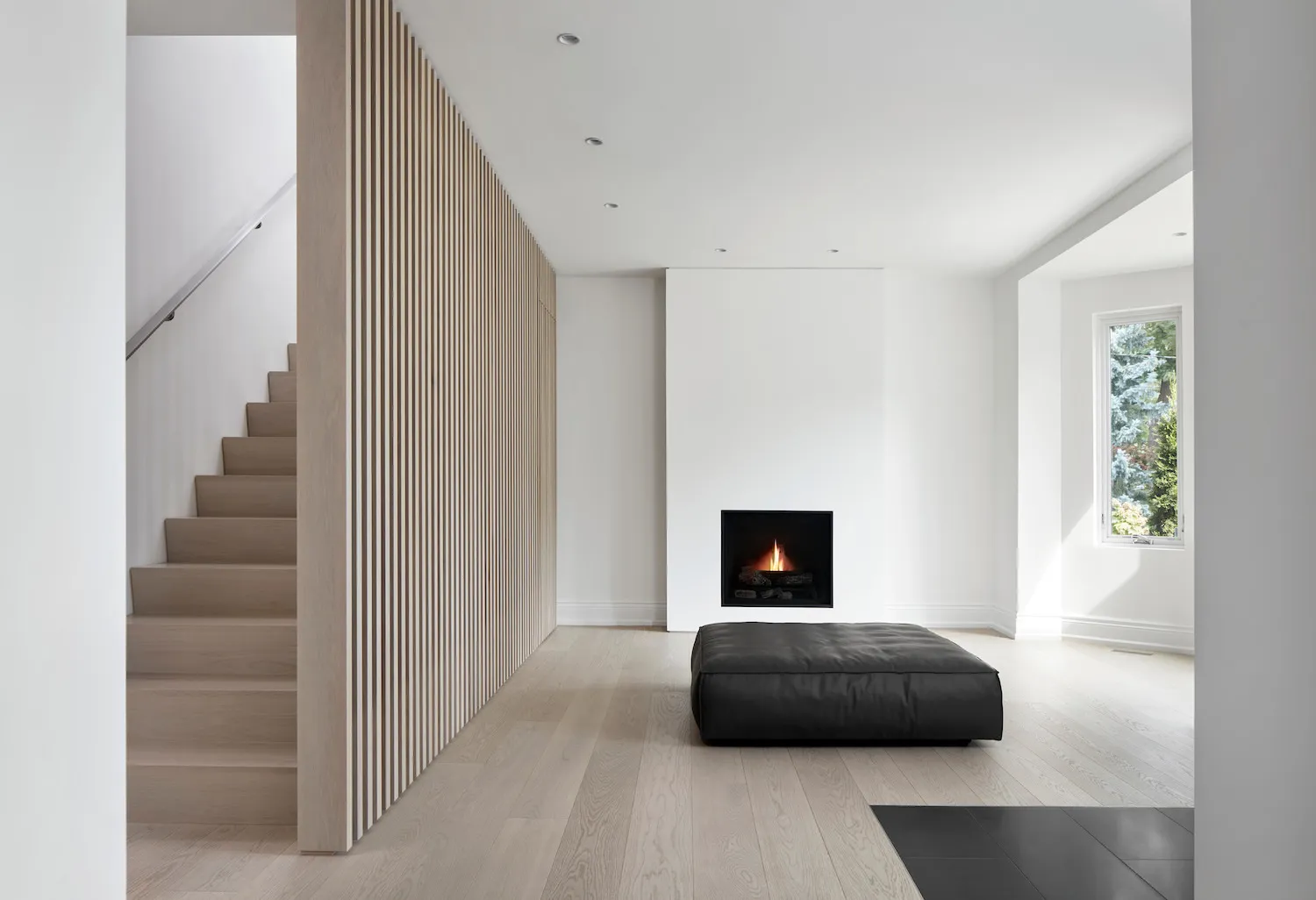 A minimalist living room featuring a light wood floor, a small black ottoman in front of a fireplace, and a white wall.