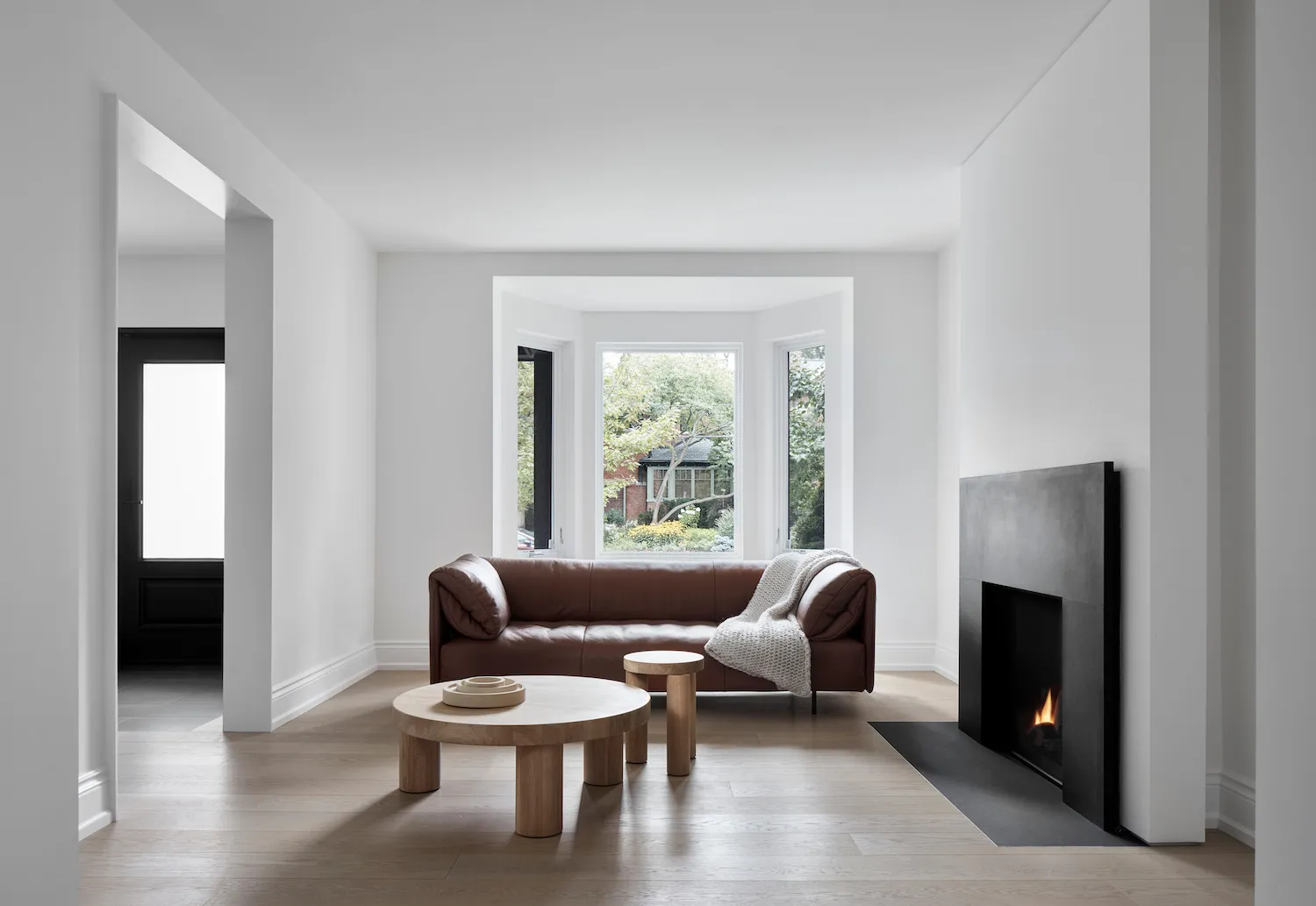 A minimalist living room featuring a brown leather sofa, wooden coffee tables, and a modern fireplace with a cozy blanket.