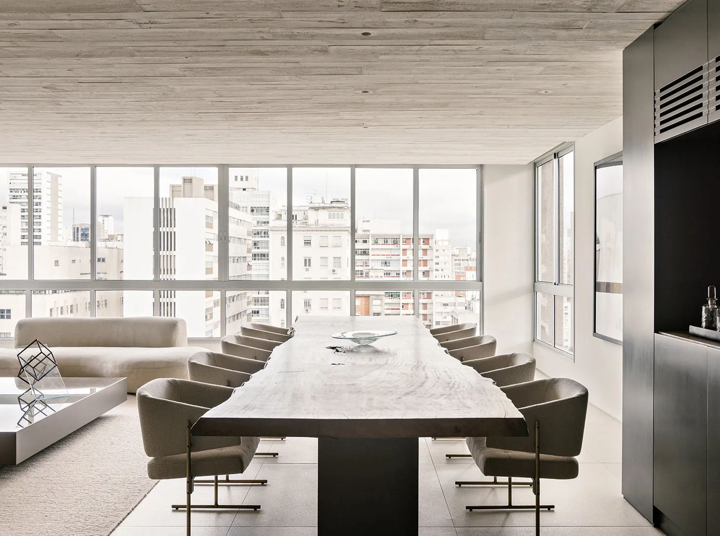 A modern conference room featuring a long wooden table surrounded by upholstered chairs and a large window with a cityscape v