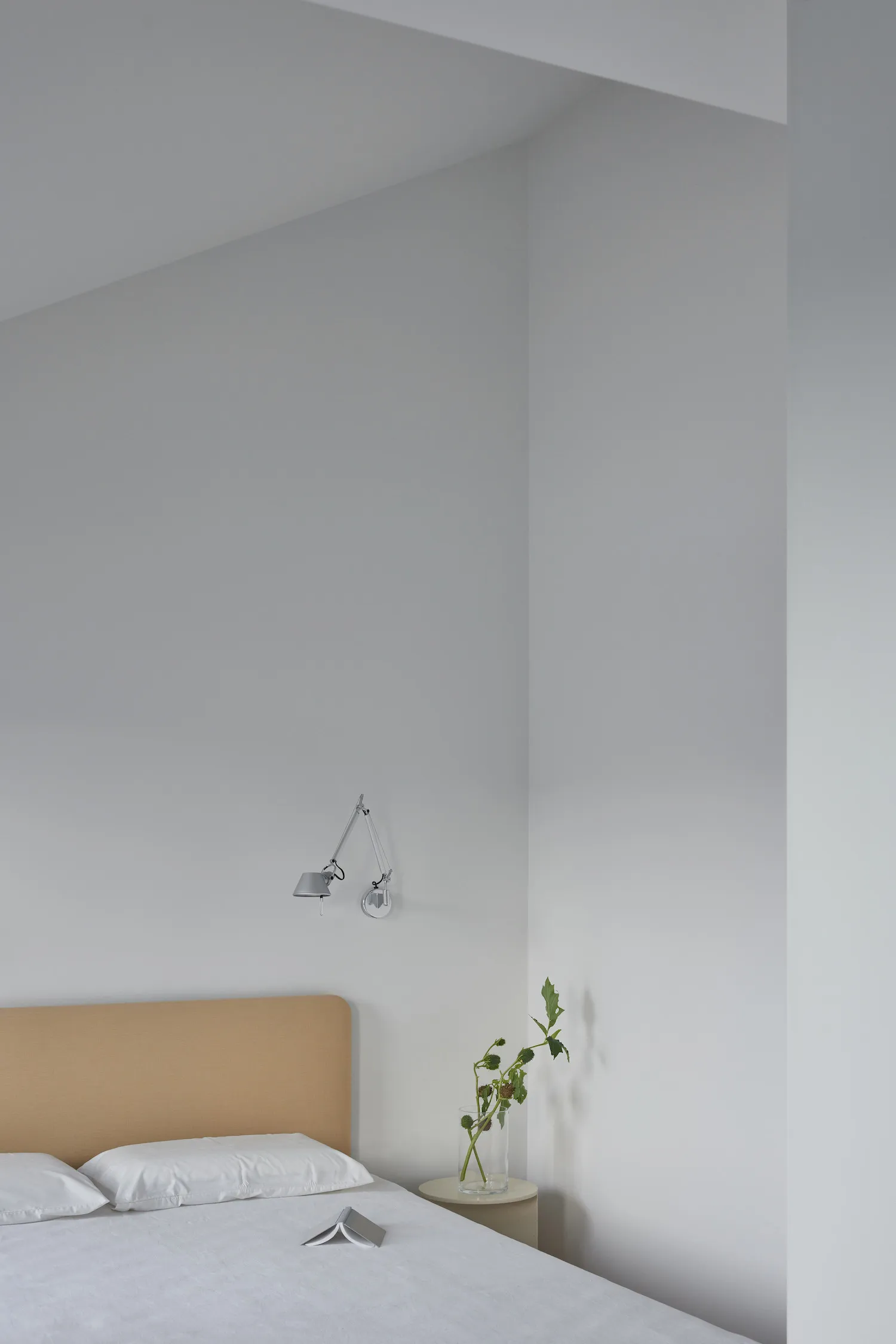 A modern minimalist bedroom featuring a bed with a fabric headboard and white bedding, placed in front of a white wall with a