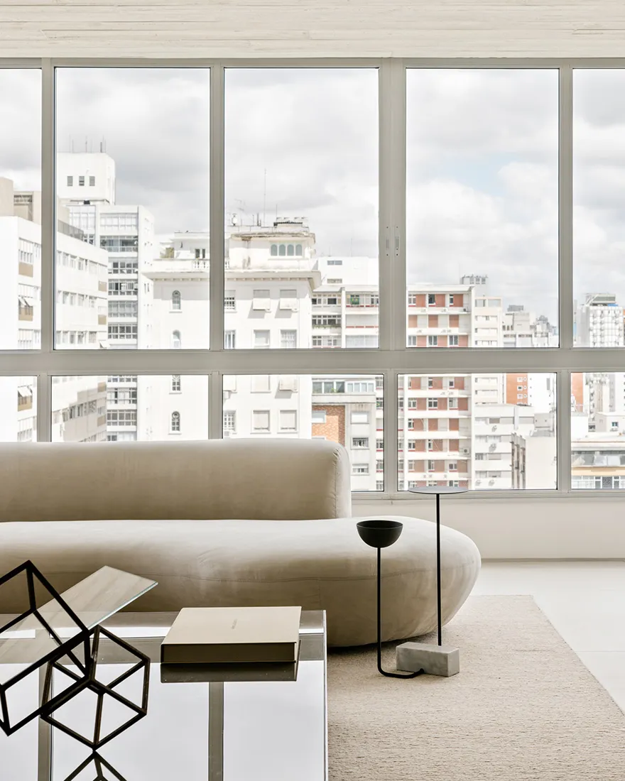 A minimalist living room with a white sofa, coffee table, and cityscape view through large windows.