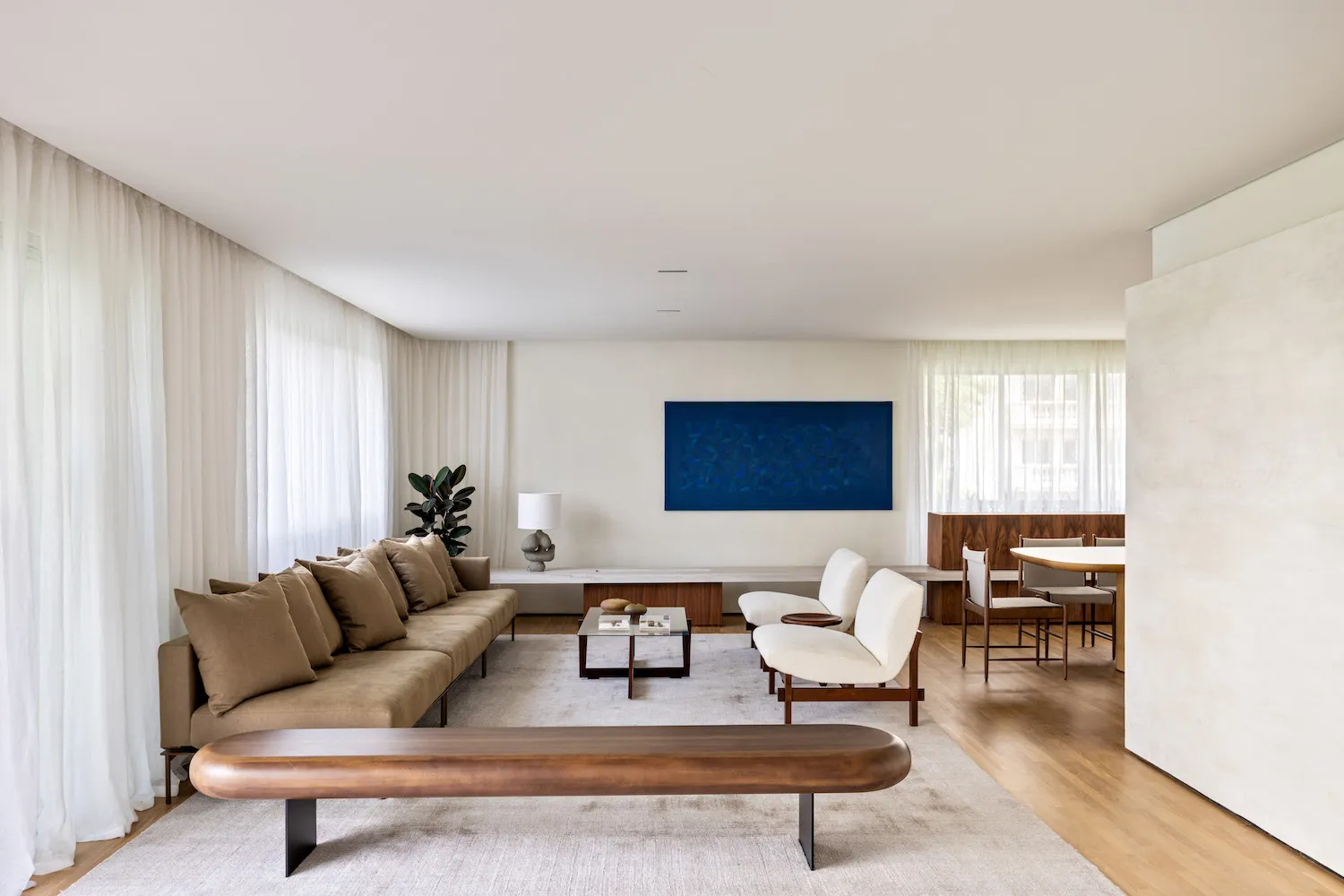 A modern living room with a minimalist design, featuring a brown sofa, white chairs, a wooden coffee table, and a blue artwor