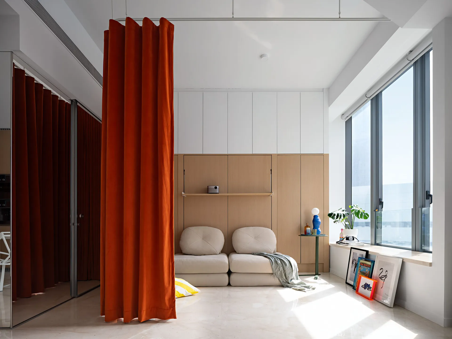 A minimalist living room with a large window, orange curtains, and a beige sofa.