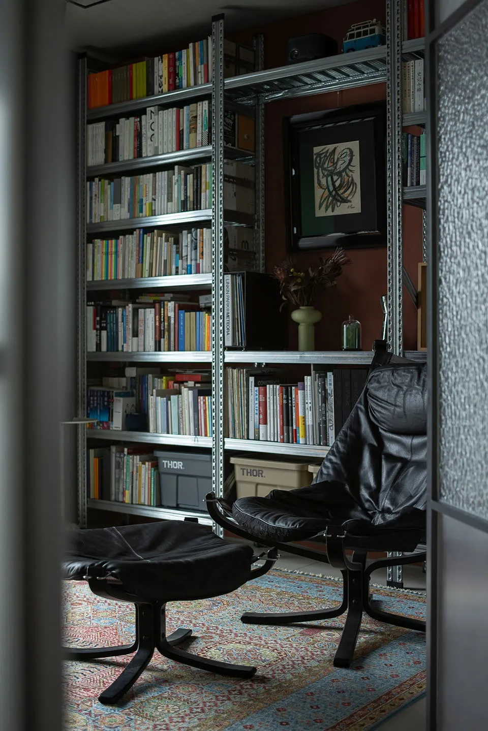 A modern library featuring metal shelves filled with books, a black leather chair and ottoman, and a patterned rug.