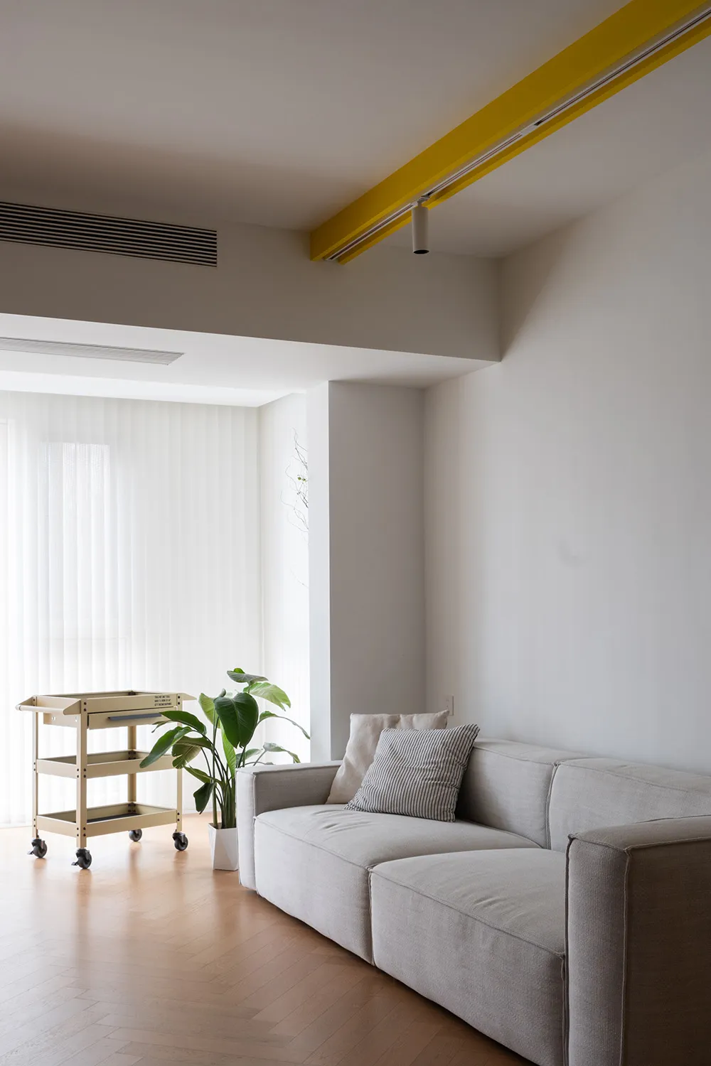 Modern minimalist living room featuring a fabric couch with a yellow track light overhead and a rolling cart with a plant.