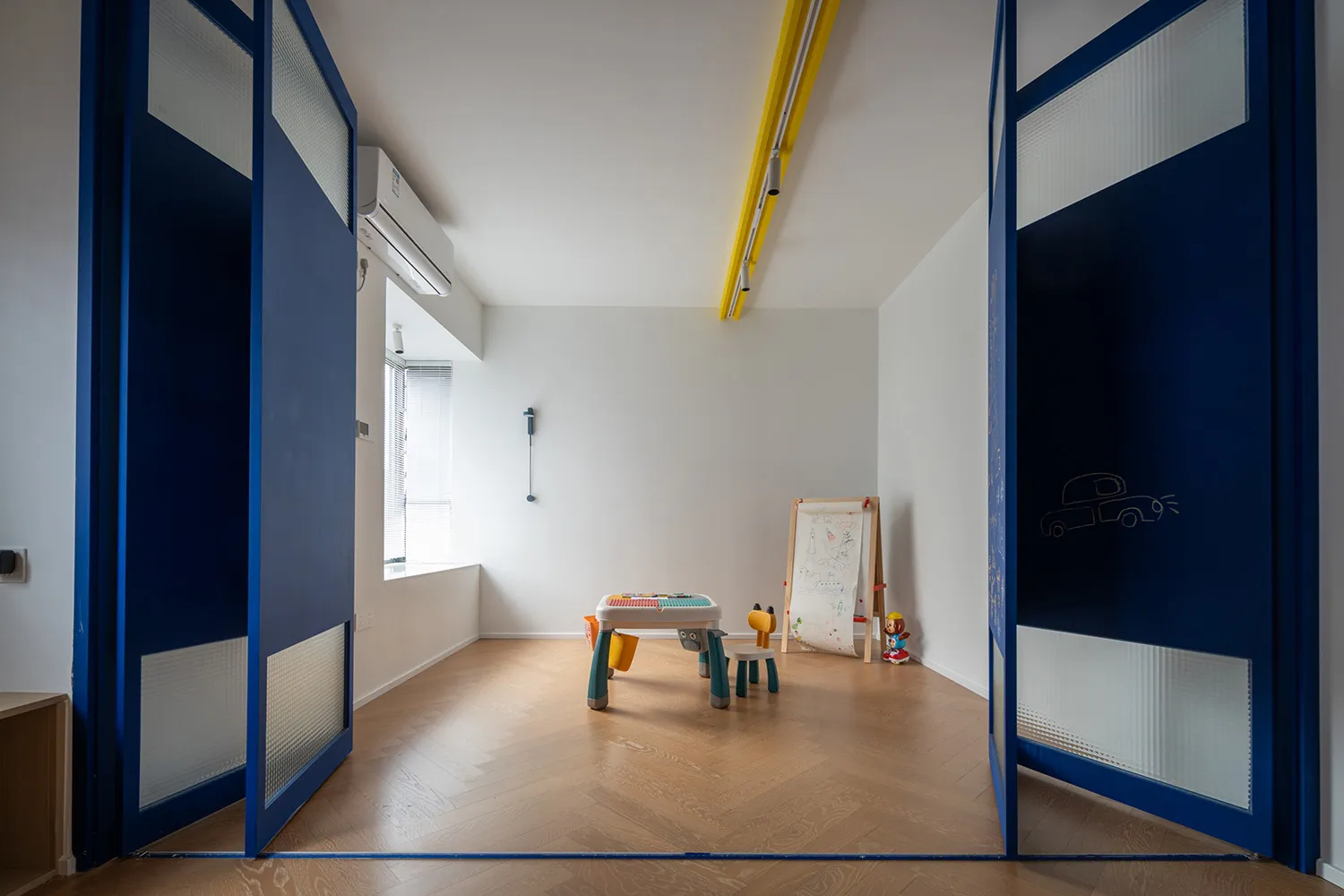 A modern children's playroom with blue double doors and wooden flooring, featuring a play table and easel.