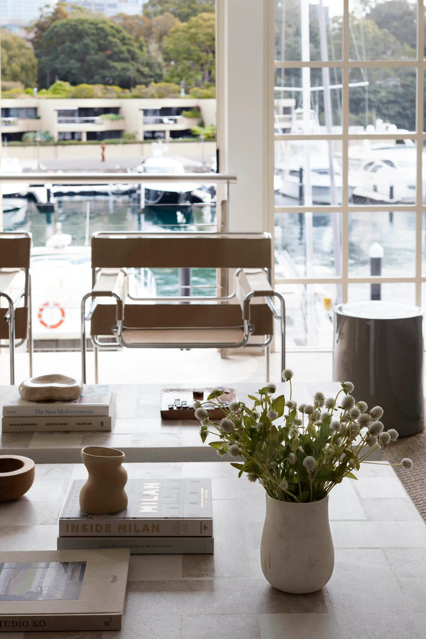A modern living room featuring wooden furniture, books, and a vase of flowers with white blooms on a table by a large window