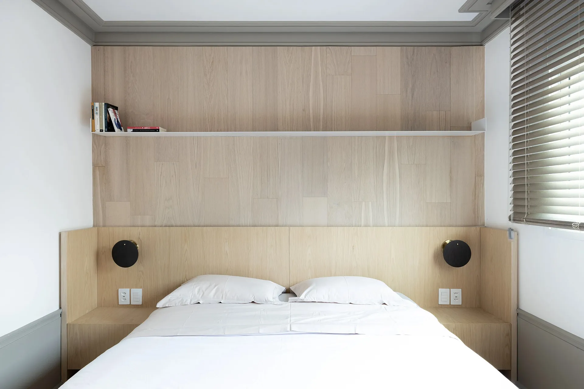 A minimalistic bedroom with wooden walls and a white bed, featuring a built-in shelf above the headboard.