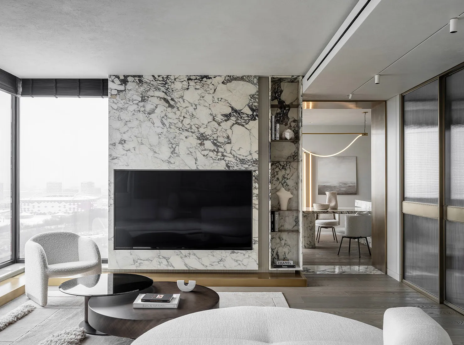 A modern living room featuring a marble accent wall with a mounted television, a round glass and wood coffee table, and a plu