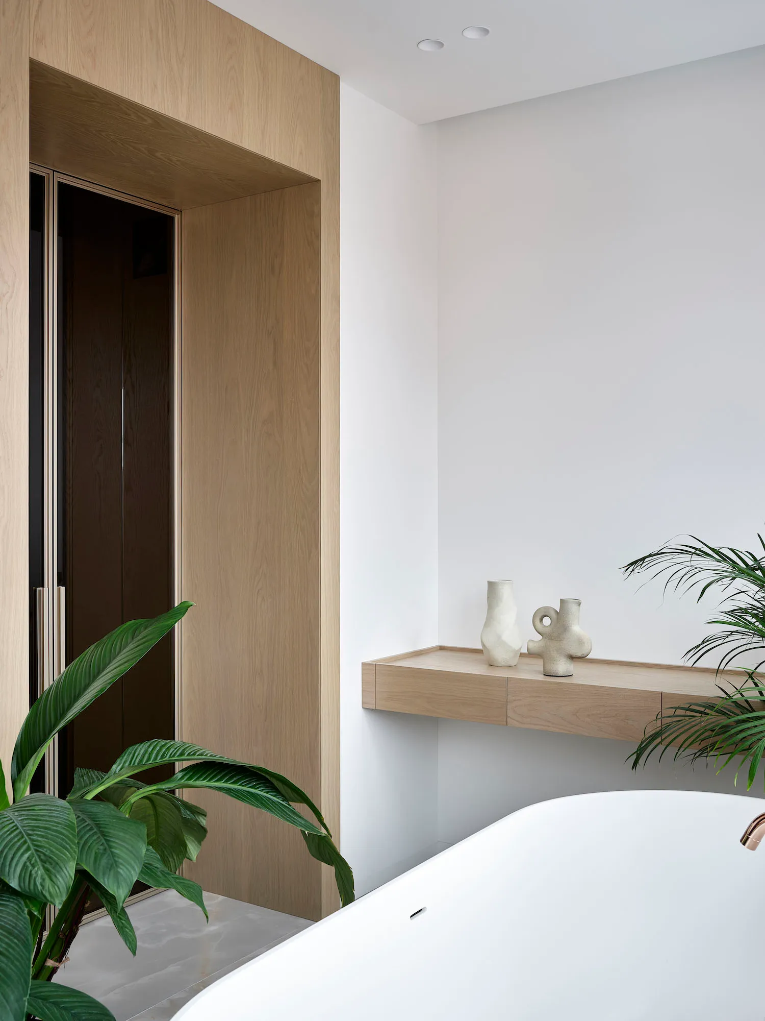 A minimalist bathroom features a white porcelain bathtub, wood shelving with decorative vases, and concrete flooring.