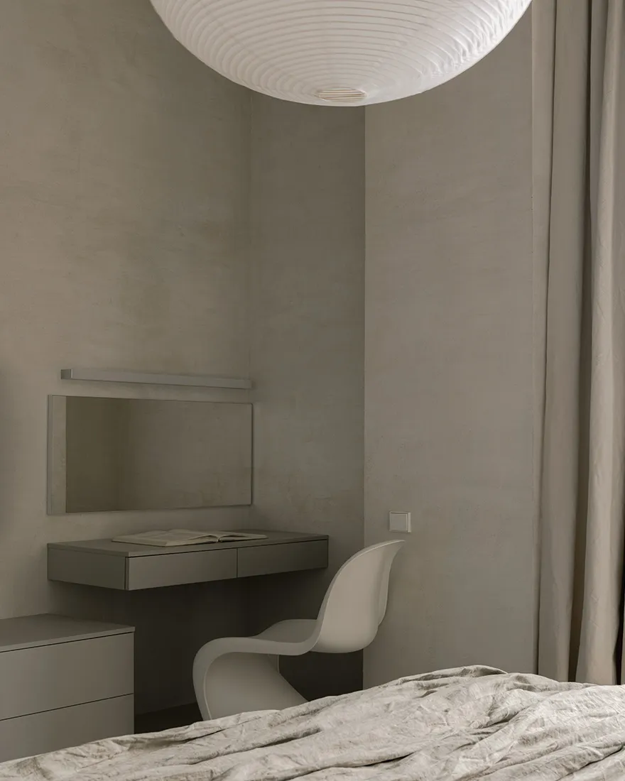 A minimalist bedroom with a bed, chair, desk, and mirror against a textured gray wall.