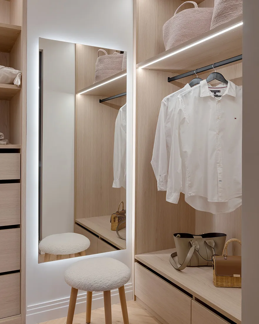 A minimalist wardrobe with wooden materials, featuring a full-length mirror, white stools, and neatly arranged clothing and a