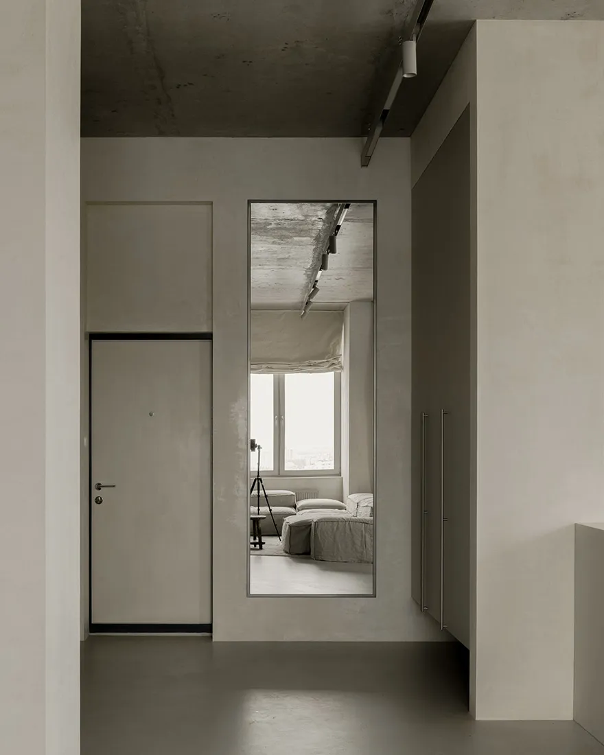 A minimalist living room with concrete walls and floor, featuring a large mirror reflecting the interior.