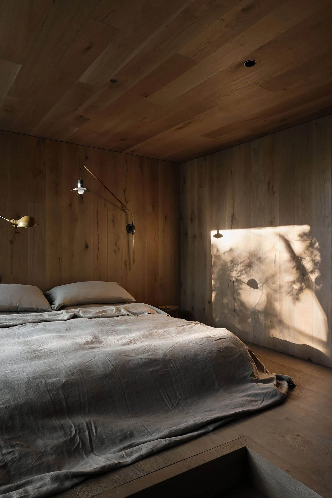 A minimalist bedroom with wooden walls and a bed with grey sheets, illuminated by natural light casting shadows on the wall.