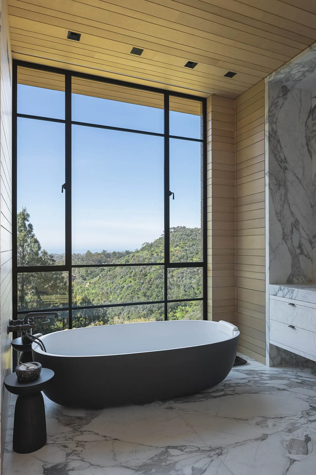 Modern bathroom featuring a freestanding bathtub with a view of the mountains, surrounded by marble and concrete materials.