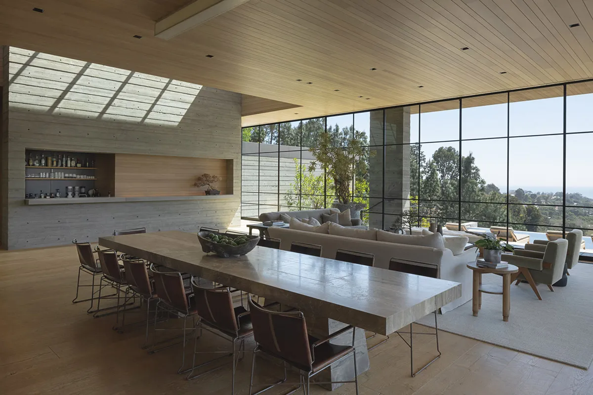 A modern living room featuring a long wooden dining table surrounded by chairs, with large windows overlooking a scenic outdo