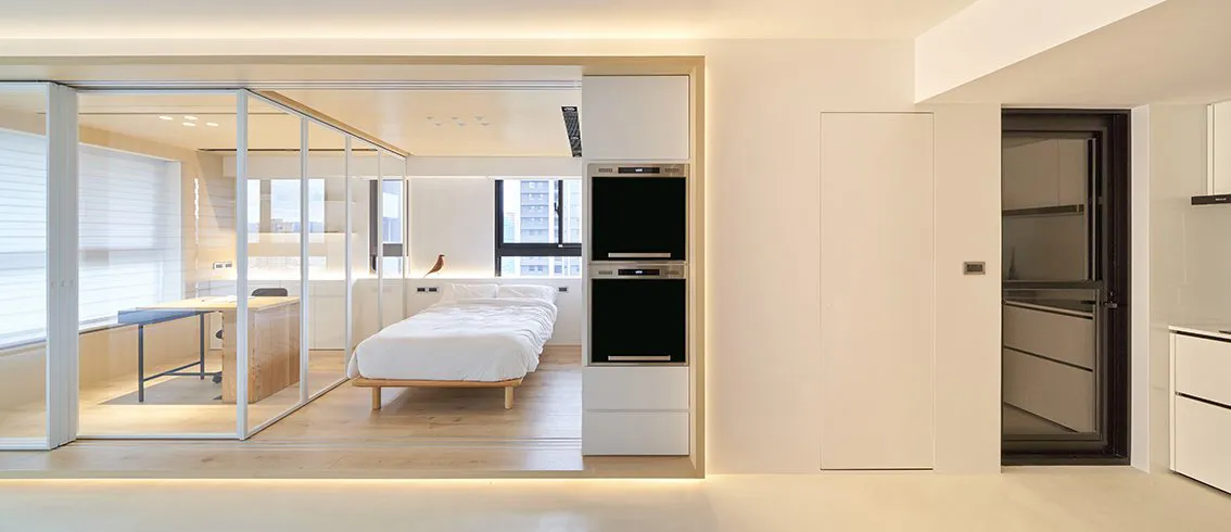 A modern, minimalist kitchen with white cabinets, a long wooden table, and a view of a bed and cityscape outside.