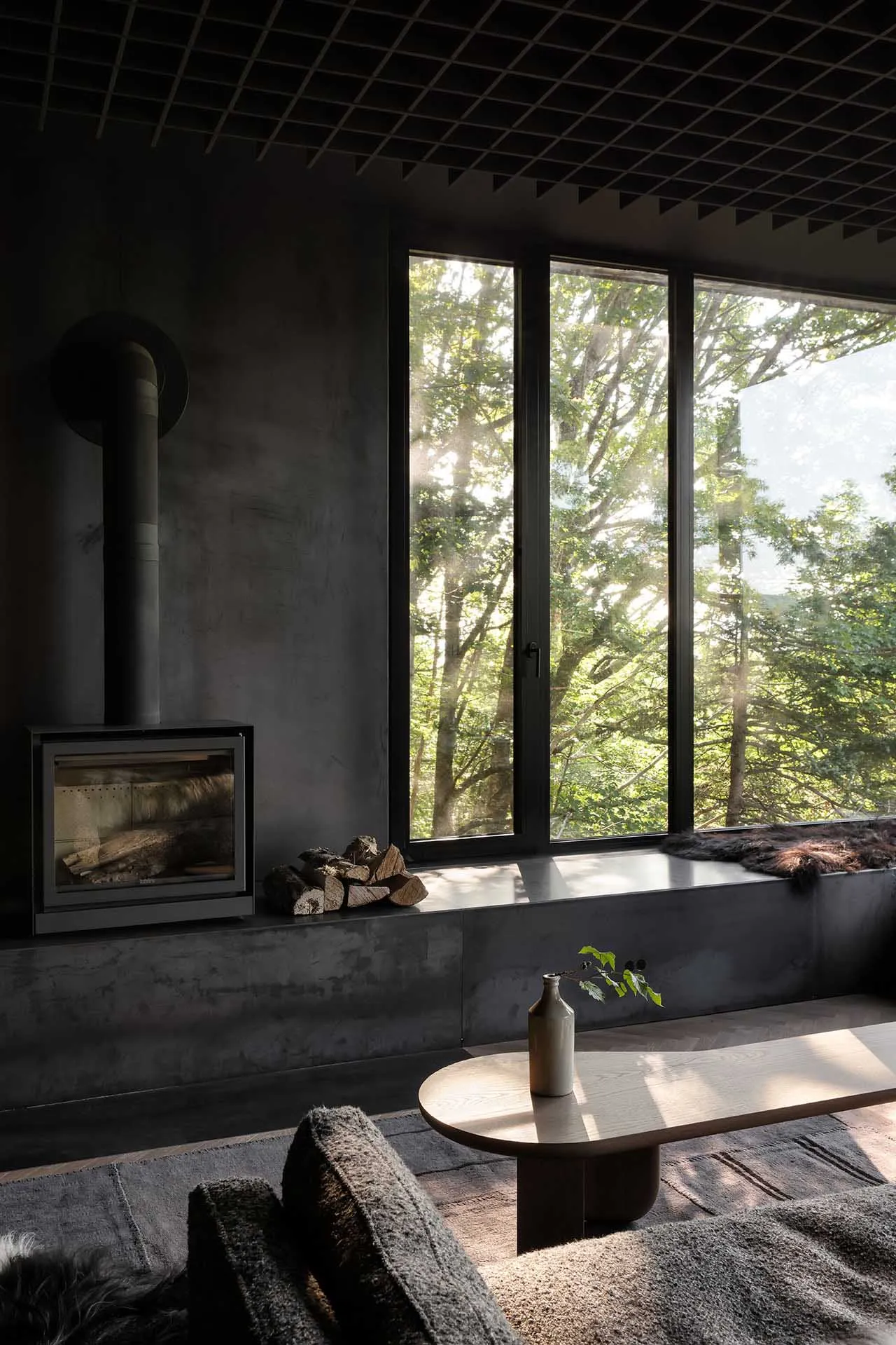 Modern living room with concrete walls and floors, featuring a fireplace and large windows overlooking a forest.