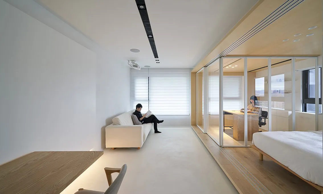A modern living room with a white sofa and wooden flooring, separated from an office space with glass walls.