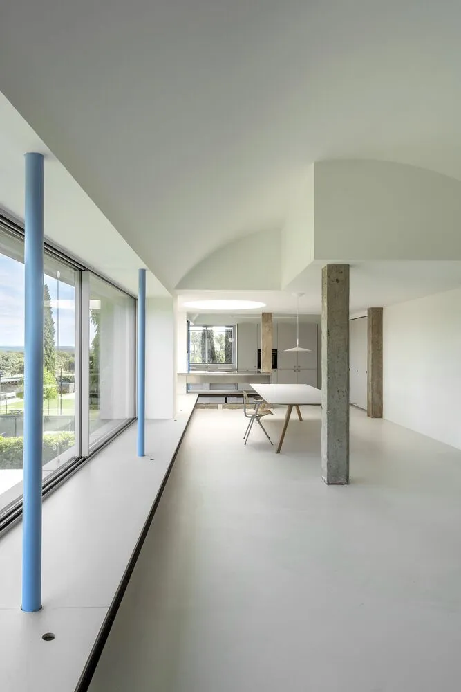 Modern minimalist living room with concrete walls and floor, large windows, and simple furniture.