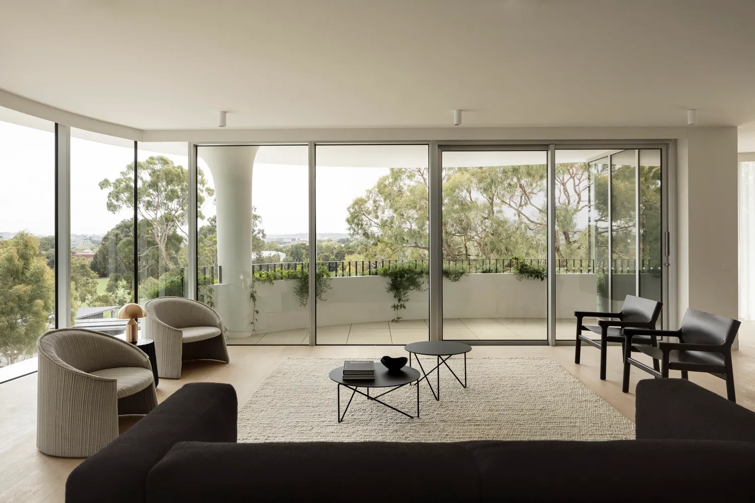 Modern living room with minimalist furniture, large windows, and a balcony view of trees.