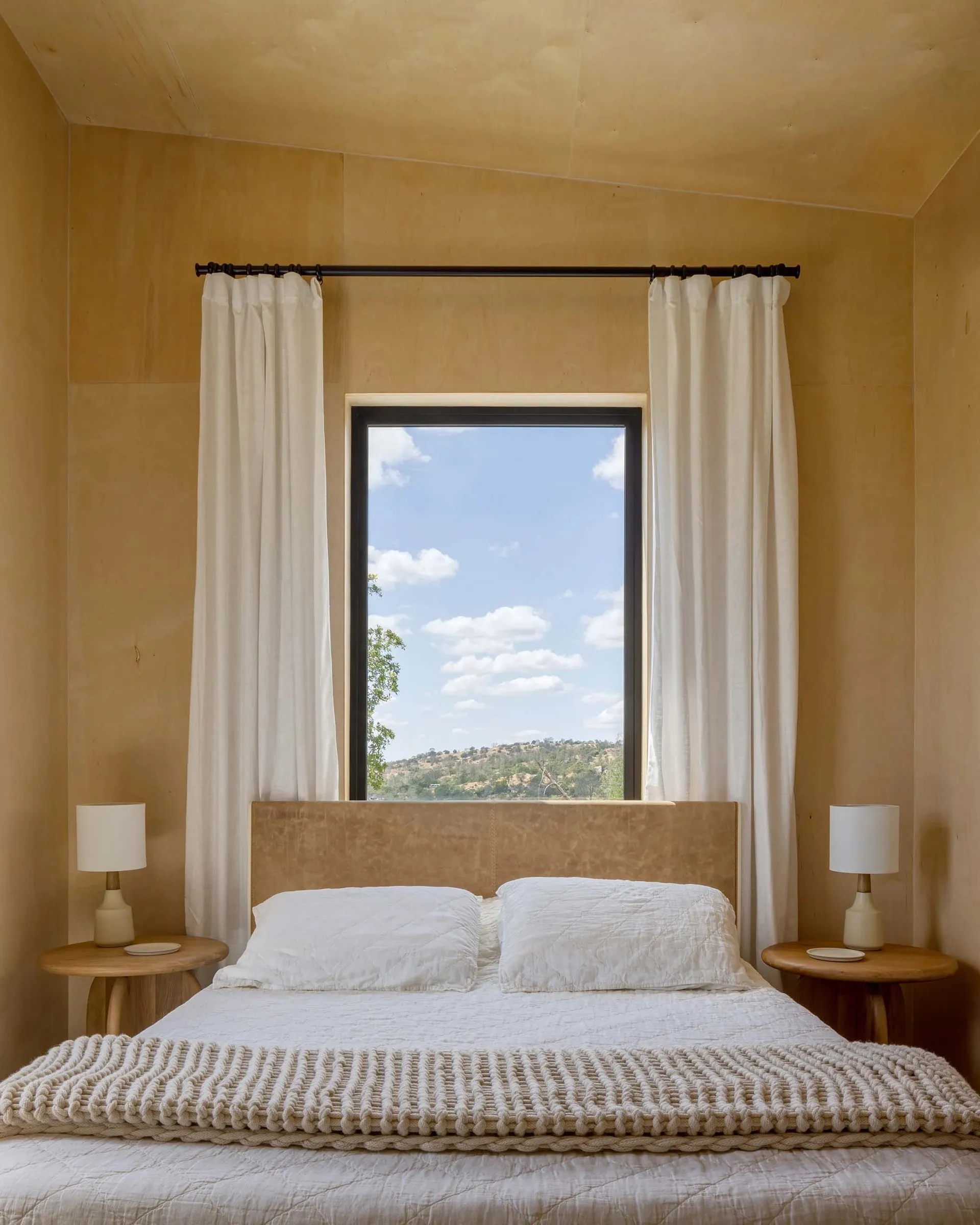 A minimalist bedroom with white curtains, a bed, and two side tables with lamps.