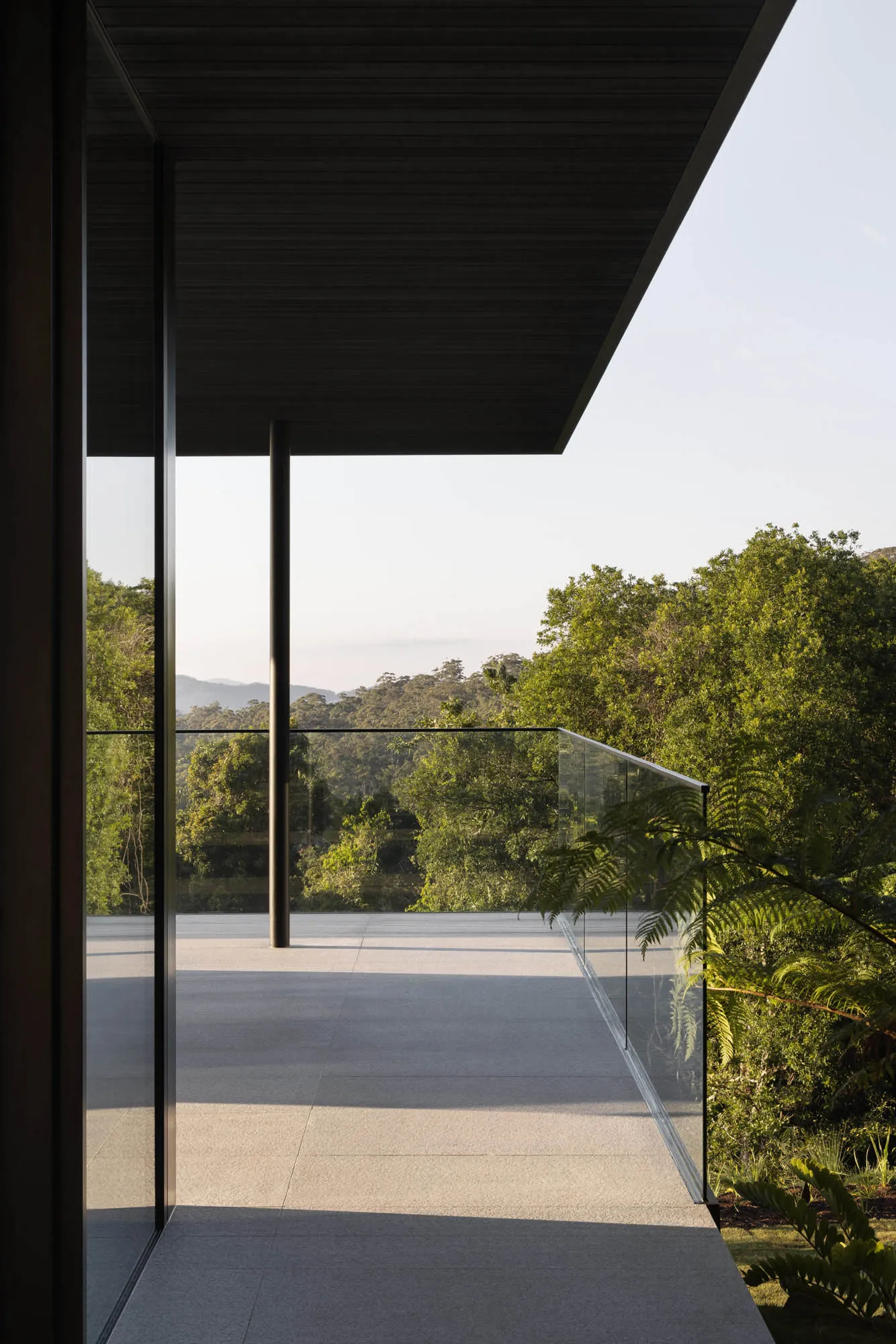 A modern residential patio with glass walls and a metal railing overlooking lush greenery.