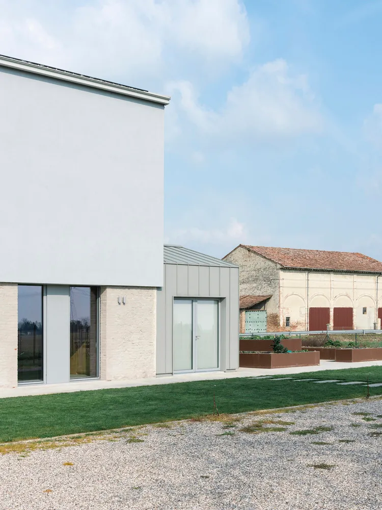 A modern exterior of a building with white concrete walls, large glass windows, and a gravel driveway with green grass.