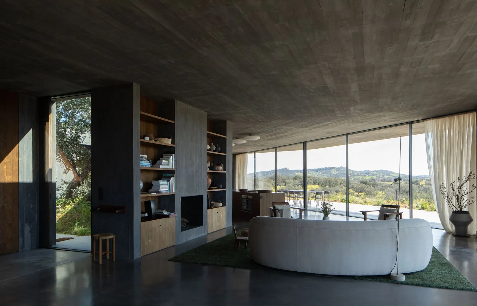Modern living room with concrete walls, floor, and ceiling, featuring a large circular sofa and a bookshelf filled with books