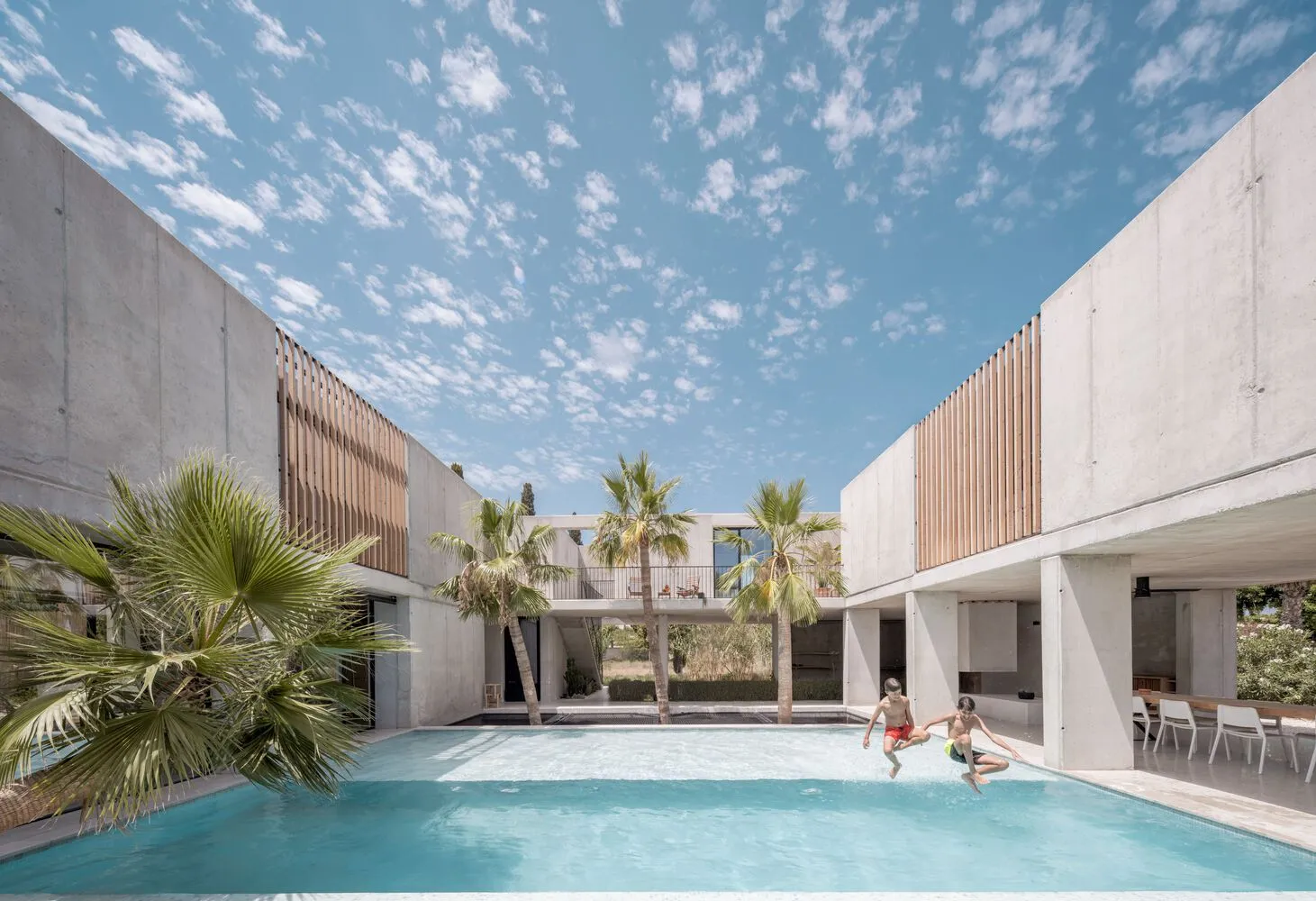 Two people jumping into a swimming pool surrounded by concrete walls with large windows and palm trees.
