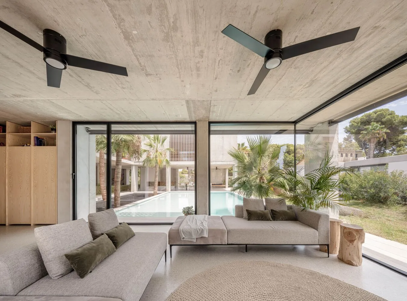 A modern living room featuring a sleek concrete ceiling and walls, with large windows offering a view of a pool and tropical 