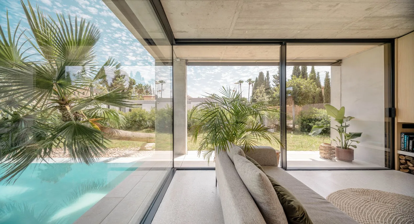 Modern living room with concrete walls and large glass window looking out to an outdoor view of palm trees and a swimming poo