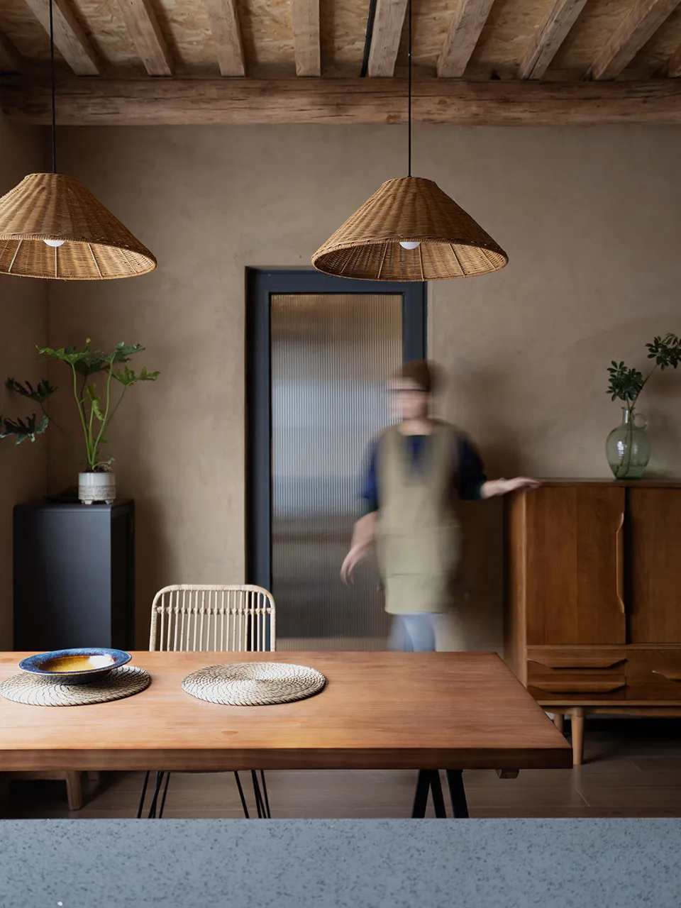 A rustic dining room featuring a wooden table with woven placemats, a concrete floor, and wicker pendant lights hanging above