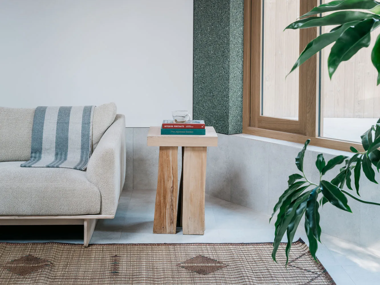 A minimalist living room featuring wooden furniture, a beige sofa, a wooden side table with books and a cup, and a plant in f