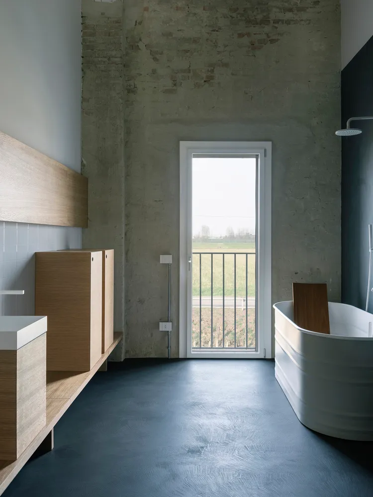 Modern bathroom with industrial design featuring concrete floor, wooden vanity, and white bathtub with a view of green fields
