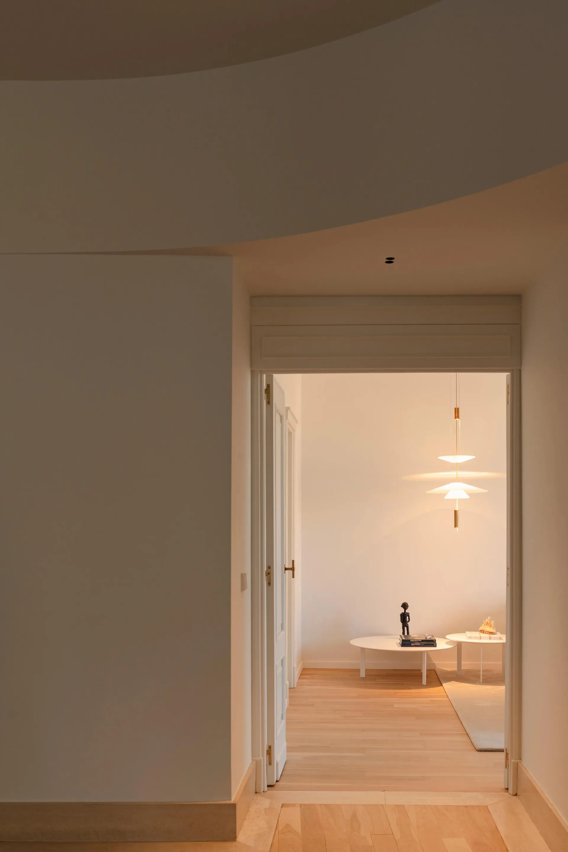 Contemporary living room with minimalistic design, featuring light-colored wood flooring, white walls, and a hanging lamp.